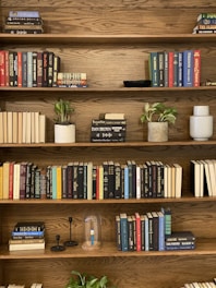 A warm-toned solid wood bookshelf with multiple tiers filled with colorful books and small plants.