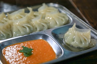 A metal tray containing neatly arranged steamed dumplings and a serving of bright orange dipping sauce garnished with a small piece of green cilantro. The dumplings have a glossy, delicate appearance with pleated edges indicating careful preparation.