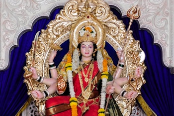A richly adorned statue of a deity is depicted, seated within an ornate gold frame. The deity has multiple arms holding various symbolic objects like a trident and a sword. The figure is dressed in vibrant red and green garments, adorned with intricate jewelry and garlands of flowers. The background is a deep purple fabric, enhancing the opulence of the scene.