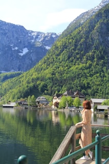 A serene lake reflecting lush green mountains and partially snow-covered peaks in the background. A quaint village with charming houses sits by the water's edge. A person in a floral dress is gazing at the scenery while standing on a wooden deck.