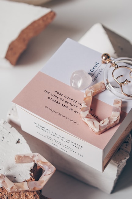 A beauty product box is displayed with stylish packaging. It includes elements like a circular metal accessory, a rose quartz crystal, and pink tortoiseshell-style earrings. The box features text promoting the product's natural and detoxifying properties, and it's set against a backdrop of textured materials.
