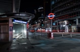 A nighttime view of an urban street corner featuring the Aldgate East Station entrance with its distinctive modern architectural canopy. The surrounding area is illuminated by the station's glowing sign and sparse streetlights. In the background, a brightly lit Underground sign is visible, along with streaks of red light from passing vehicles. The scene exhibits a contrast between the dimly lit street and the lights from nearby buildings.