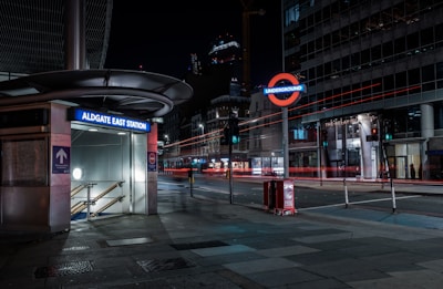 A nighttime view of an urban street corner featuring the Aldgate East Station entrance with its distinctive modern architectural canopy. The surrounding area is illuminated by the station's glowing sign and sparse streetlights. In the background, a brightly lit Underground sign is visible, along with streaks of red light from passing vehicles. The scene exhibits a contrast between the dimly lit street and the lights from nearby buildings.