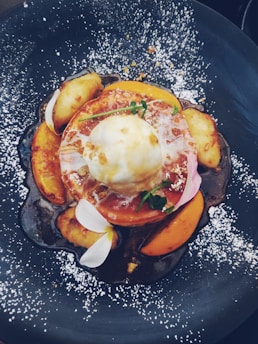 A beautifully plated dessert featuring a scoop of ice cream on top of caramelized fruit slices, garnished with edible flowers and a sprig of greenery. Powdered sugar is sprinkled across the dark plate, creating an artistic contrast.