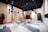 The image depicts a spacious and well-organized yoga studio. The room has several aerial yoga fabrics suspended from the ceiling, which are turquoise in color. There are yoga mats evenly spaced on the floor, each with a foam block placed on top. The studio features a light wooden floor, light-colored walls, and a dark ceiling with lights. One wall has several hooks with more aerial yoga fabrics, and there are potted plants adding an element of nature to the room. At the end of the room, there is a storage area with shelves and more equipment.