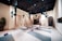 The image depicts a spacious and well-organized yoga studio. The room has several aerial yoga fabrics suspended from the ceiling, which are turquoise in color. There are yoga mats evenly spaced on the floor, each with a foam block placed on top. The studio features a light wooden floor, light-colored walls, and a dark ceiling with lights. One wall has several hooks with more aerial yoga fabrics, and there are potted plants adding an element of nature to the room. At the end of the room, there is a storage area with shelves and more equipment.