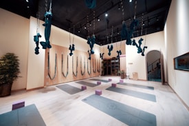 The image depicts a spacious and well-organized yoga studio. The room has several aerial yoga fabrics suspended from the ceiling, which are turquoise in color. There are yoga mats evenly spaced on the floor, each with a foam block placed on top. The studio features a light wooden floor, light-colored walls, and a dark ceiling with lights. One wall has several hooks with more aerial yoga fabrics, and there are potted plants adding an element of nature to the room. At the end of the room, there is a storage area with shelves and more equipment.