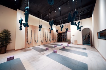 The image depicts a spacious and well-organized yoga studio. The room has several aerial yoga fabrics suspended from the ceiling, which are turquoise in color. There are yoga mats evenly spaced on the floor, each with a foam block placed on top. The studio features a light wooden floor, light-colored walls, and a dark ceiling with lights. One wall has several hooks with more aerial yoga fabrics, and there are potted plants adding an element of nature to the room. At the end of the room, there is a storage area with shelves and more equipment.