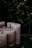 A group of large, unlit candles with a textured surface arranged on a dark tray. They are set against a backdrop of lush, green foliage.