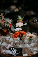A collection of decorative glass vases arranged on a circular tray, interspersed with vibrant flowers including orange marigolds and pale pink roses. The setup includes a small chalkboard sign displaying a price. The surrounding is dimly lit, which enhances the focus on the floral arrangement.