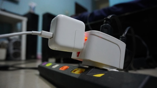 A close-up view of a power strip with multiple electronic devices plugged into it. The focus is on a white charging adapter with a glowing orange light connected to a USB cable. The background is slightly blurred, showing a room with various electronic equipment.