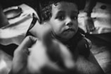A black and white photo featuring a young child with large, expressive eyes looking towards the camera. The child is holding a camera lens cap labeled 'Sony' in their hands. There are blurred hands in the foreground, possibly indicating movement or reaching towards the child. The setting appears to be indoors with a visible cloth-covered surface in the background.