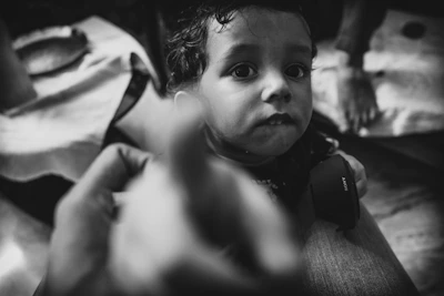 A black and white photo featuring a young child with large, expressive eyes looking towards the camera. The child is holding a camera lens cap labeled 'Sony' in their hands. There are blurred hands in the foreground, possibly indicating movement or reaching towards the child. The setting appears to be indoors with a visible cloth-covered surface in the background.