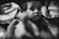 A black and white photo featuring a young child with large, expressive eyes looking towards the camera. The child is holding a camera lens cap labeled 'Sony' in their hands. There are blurred hands in the foreground, possibly indicating movement or reaching towards the child. The setting appears to be indoors with a visible cloth-covered surface in the background.