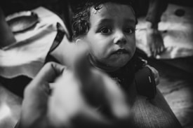 A black and white photo featuring a young child with large, expressive eyes looking towards the camera. The child is holding a camera lens cap labeled 'Sony' in their hands. There are blurred hands in the foreground, possibly indicating movement or reaching towards the child. The setting appears to be indoors with a visible cloth-covered surface in the background.