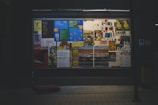 A window display showcases various colorful posters arranged closely together. The posters display a mix of text and images, with some in Cyrillic script. The overall setting seems urban and slightly dimly lit, with the illuminated posters providing contrast. The background is dark, emphasizing the vibrancy of the posters.