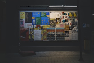 A window display showcases various colorful posters arranged closely together. The posters display a mix of text and images, with some in Cyrillic script. The overall setting seems urban and slightly dimly lit, with the illuminated posters providing contrast. The background is dark, emphasizing the vibrancy of the posters.
