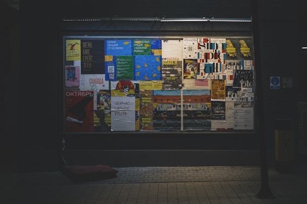 A window display showcases various colorful posters arranged closely together. The posters display a mix of text and images, with some in Cyrillic script. The overall setting seems urban and slightly dimly lit, with the illuminated posters providing contrast. The background is dark, emphasizing the vibrancy of the posters.