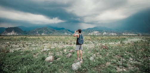 A person with a backpack stands on a rocky terrain, capturing a photograph of the distant mountains under a cloudy sky. The landscape is a mix of grass, rocks, and sparse vegetation, with mountains and dark clouds creating a dramatic backdrop.
