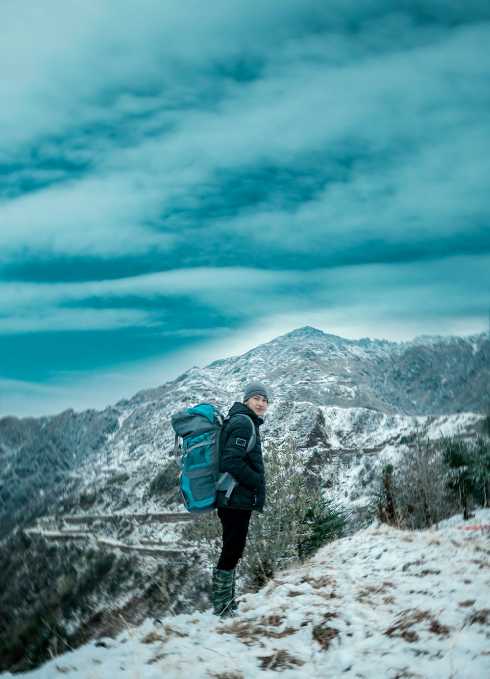 Hombre con chaqueta negra y mochila azul de pie en el suelo cubierto de nieve durante el día