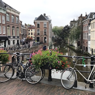 A picturesque European canal scene featuring a bridge adorned with vibrant flowers and bicycles resting against a railing. The background showcases traditional brick buildings lining the canal, with lush greenery and trees complementing the serene waterway. The cobblestone path and classic architecture add to the charm and elegance of the setting.