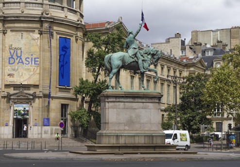 A statue of a person on horseback is situated prominently in front of a historic building. The statue features a person holding a flag, and it is crafted in a dark patina. Behind the statue, the building has large banners and ornate architectural details. There are trees and urban elements such as a street sign and parked vehicles nearby.