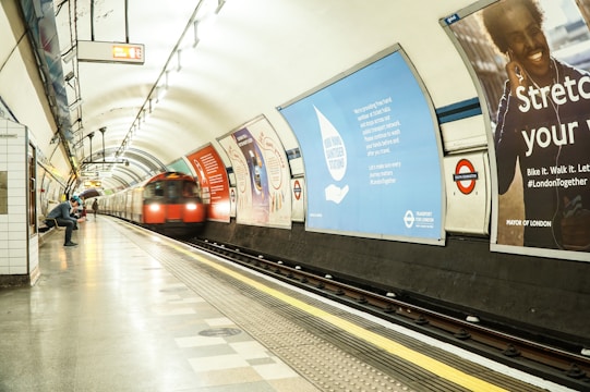 A subterranean metro station with a sleek, curved ceiling and bright fluorescent lighting. The platform is mostly empty, except for a few individuals sitting and standing. A red underground train zooms by on the tracks, creating a blur of motion. Various advertisements line the walls of the station, with messages related to transportation and community initiatives.