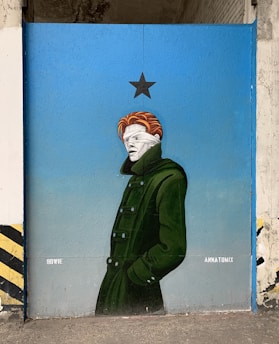A mural depicts a figure with its head wrapped in bandages, wearing a dark green trench coat. The figure stands against a backdrop of a blue wall with a black star above its head. The artist's name, ANKATOMIX, is visible on the right side of the mural and the name BOWIE is on the left.
