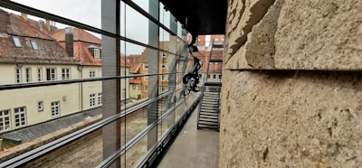 A modern architectural perspective features a narrow passageway between a stone wall on the right and a glass window facade on the left, offering a view of traditional European-style buildings with tiled roofs in the background. An abstract black sculpture hangs from the ceiling near the window frame.