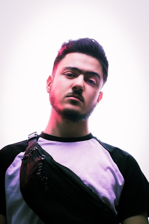 A young man with a well-groomed beard and styled hair is looking directly at the camera. He wears a t-shirt with black sleeves and a white torso, and a black cross-body bag. The lighting creates a pinkish hue on one side of his face, giving a moody and artistic effect.