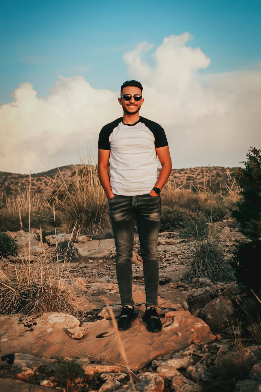 man in white crew neck t-shirt and black pants standing on rocky ground under cloudy
