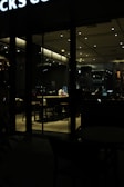 A dimly lit coffee shop interior is visible through the glass. A person is sitting alone at a table, engrossed in their work on a laptop. The ambiance is calm and quiet, with soft lighting illuminating the surroundings. Tables and bar stools are arranged neatly, reflecting a tidy and organized space. The exterior signage partially visible at the top suggests a popular chain coffee shop setting.