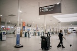 A largely empty airport terminal with rows of self-service kiosks. Overhead signage indicates directions to international arrivals and check-in areas. A person is cleaning the floor near a cart loaded with cleaning supplies. The atmosphere is quiet and unhurried.