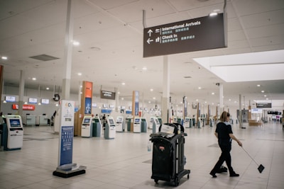 A largely empty airport terminal with rows of self-service kiosks. Overhead signage indicates directions to international arrivals and check-in areas. A person is cleaning the floor near a cart loaded with cleaning supplies. The atmosphere is quiet and unhurried.
