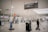 A largely empty airport terminal with rows of self-service kiosks. Overhead signage indicates directions to international arrivals and check-in areas. A person is cleaning the floor near a cart loaded with cleaning supplies. The atmosphere is quiet and unhurried.