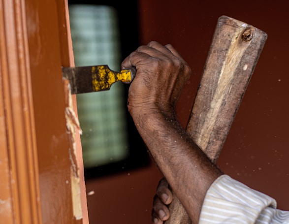 A close-up of a person's hands using a chisel and hammer to remove wood from a door frame. The visible hand is holding a yellow-handled chisel, while the other grips a worn wooden mallet. The door frame is brown, and there are visible wood shavings.