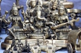 A collection of intricately designed brass statues depicting Hindu deities is arranged closely together. The central figure appears to be Ganesha, with large ears and an elephant trunk, surrounded by other detailed figures adorned with jewelry and traditional attire. The craftsmanship highlights detailed carvings and embellishments.