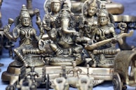 A collection of intricately designed brass statues depicting Hindu deities is arranged closely together. The central figure appears to be Ganesha, with large ears and an elephant trunk, surrounded by other detailed figures adorned with jewelry and traditional attire. The craftsmanship highlights detailed carvings and embellishments.