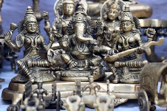 A collection of intricately designed brass statues depicting Hindu deities is arranged closely together. The central figure appears to be Ganesha, with large ears and an elephant trunk, surrounded by other detailed figures adorned with jewelry and traditional attire. The craftsmanship highlights detailed carvings and embellishments.