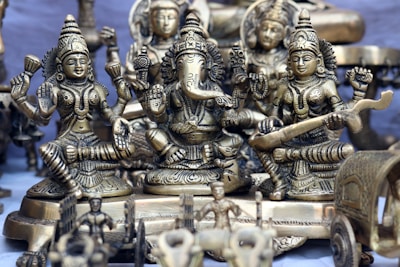 A collection of intricately designed brass statues depicting Hindu deities is arranged closely together. The central figure appears to be Ganesha, with large ears and an elephant trunk, surrounded by other detailed figures adorned with jewelry and traditional attire. The craftsmanship highlights detailed carvings and embellishments.
