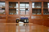 A wooden cabinet with glass doors contains various scientific instruments. In front of the cabinet, there are several brown bottles with blue and white caps placed on a wooden surface.