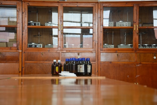 A wooden cabinet with glass doors contains various scientific instruments. In front of the cabinet, there are several brown bottles with blue and white caps placed on a wooden surface.