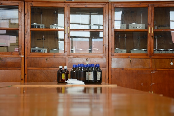 A wooden cabinet with glass doors contains various scientific instruments. In front of the cabinet, there are several brown bottles with blue and white caps placed on a wooden surface.
