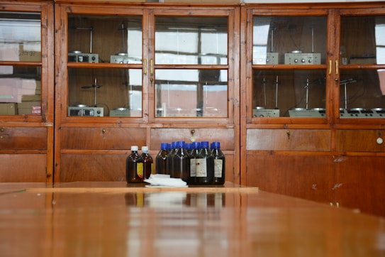 A wooden cabinet with glass doors contains various scientific instruments. In front of the cabinet, there are several brown bottles with blue and white caps placed on a wooden surface.