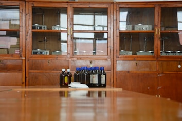 A wooden cabinet with glass doors contains various scientific instruments. In front of the cabinet, there are several brown bottles with blue and white caps placed on a wooden surface.