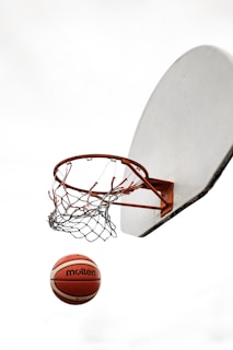 A basketball is suspended in mid-air near a net and hoop against a plain sky background. The hoop is attached to a backboard, and the image captures the dynamic movement of the ball as it approaches the basket.