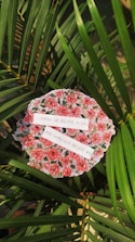 A circular fabric with a floral pattern featuring red and pink roses is placed among green palm leaves. Two strips of paper with handwritten text are laid across the fabric. The text reads: 'I KNOW WE BELIEVE IN GOD. AND I KNOW GOD BELIEVE IN US.' Sunlight is casting shadows, adding depth to the scene.