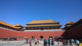 A large historic Chinese building with intricate architectural details featuring golden roofs and red walls. The foreground is bustling with people walking and gathering, suggesting a popular tourist destination. The sky is clear and bright blue.