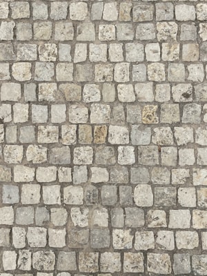 An arrangement of cobblestones forming a textured, uneven surface. The stones are rectangular and vary slightly in color, with shades of gray, beige, and hints of brown. They are laid out in a grid-like pattern, creating a rustic and historic appearance.