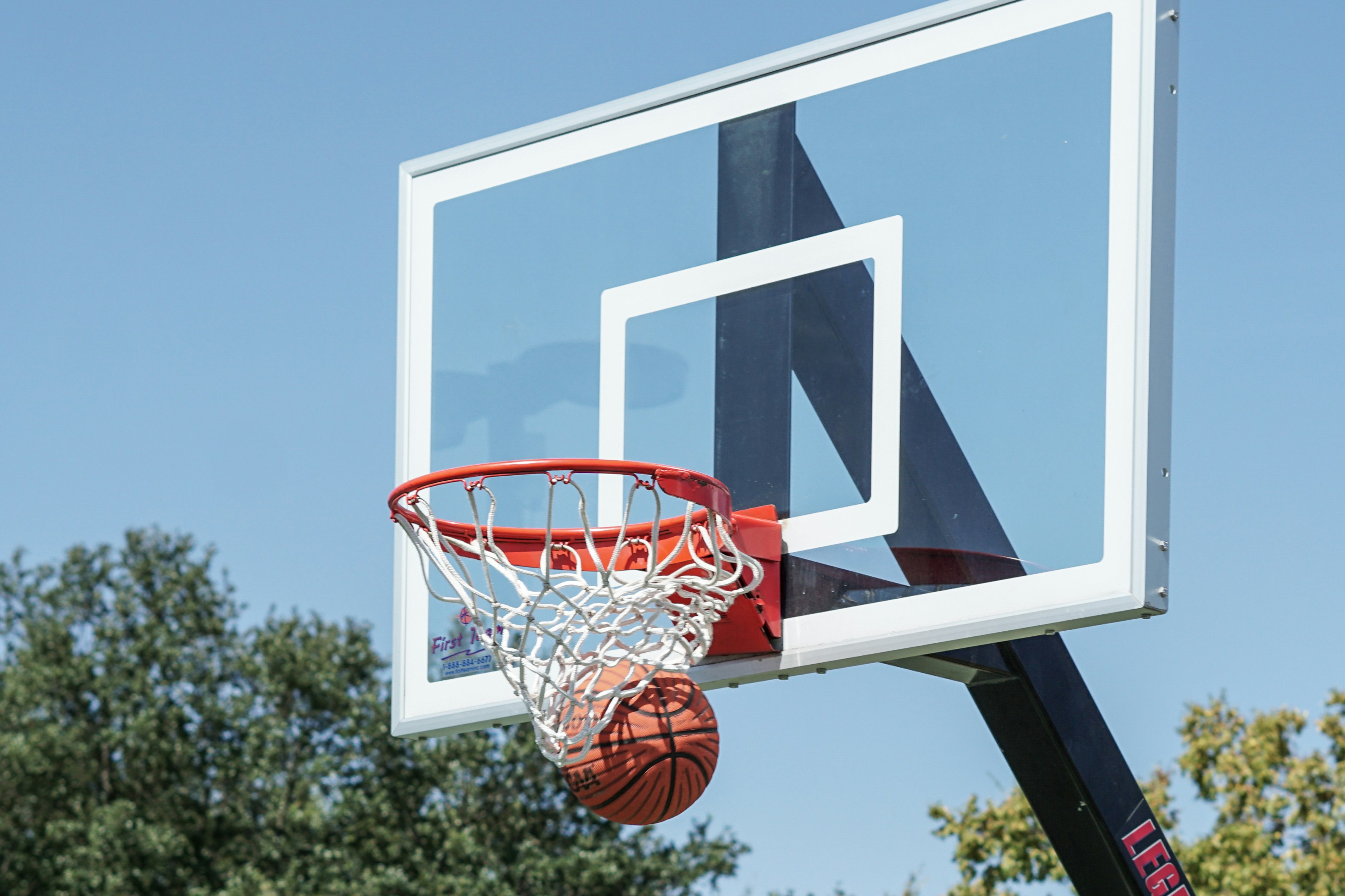 A basket ball going into a basket next to a blue sky with trees in the background.